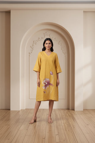 A woman modeling a mustard yellow linen tunic dress with a purple orchid print, standing in front of a beige arched wall on a wooden floor.