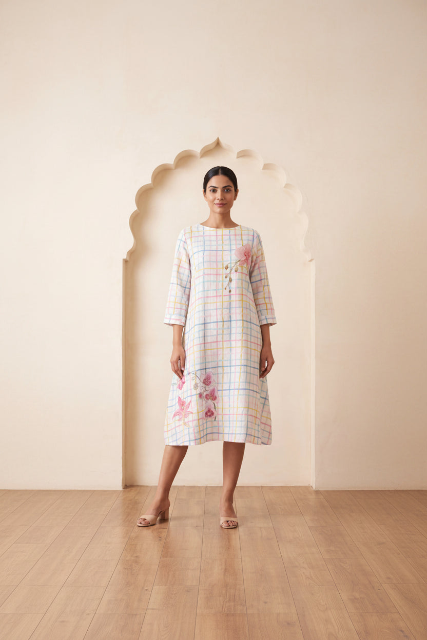 A woman models a three-quarter sleeve midi dress with a pastel multicolor plaid pattern and pink floral embroidery, standing in front of a beige wall with an arched niche.
