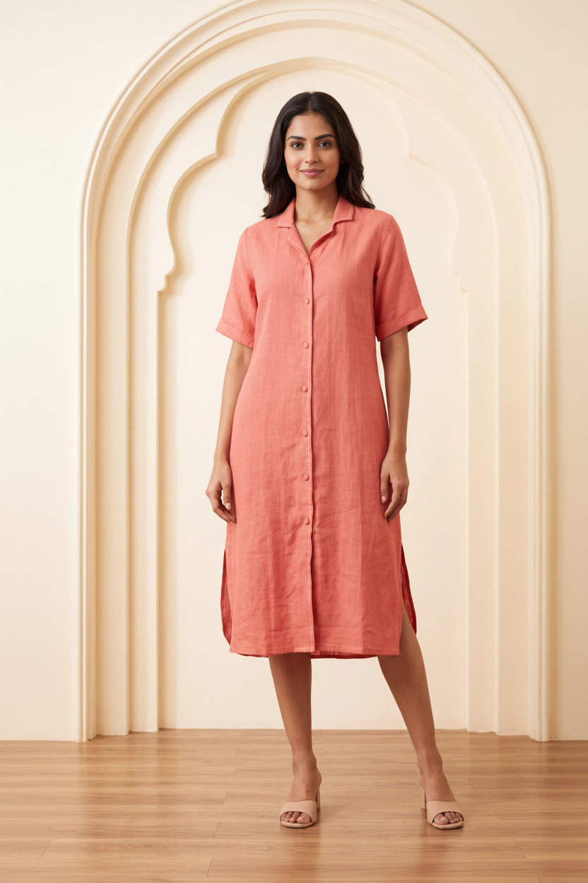 A woman modeling a short-sleeve linen shirt dress in a dusty coral pink color, standing in front of a cream-colored arched wall with a wooden floor.