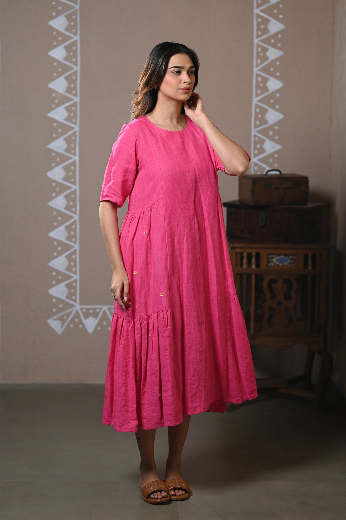 A woman wearing a bright pink tiered linen dress with short sleeves and a relaxed fit, standing in a styled room with a wooden chest and patterned wall.