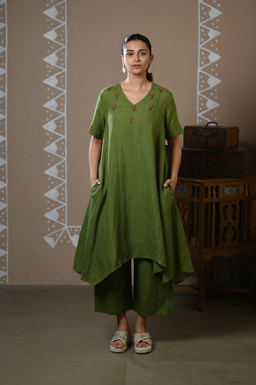 A woman modeling a green kurta and trouser set with small red embroidered details at the neckline, standing against a patterned beige wall.